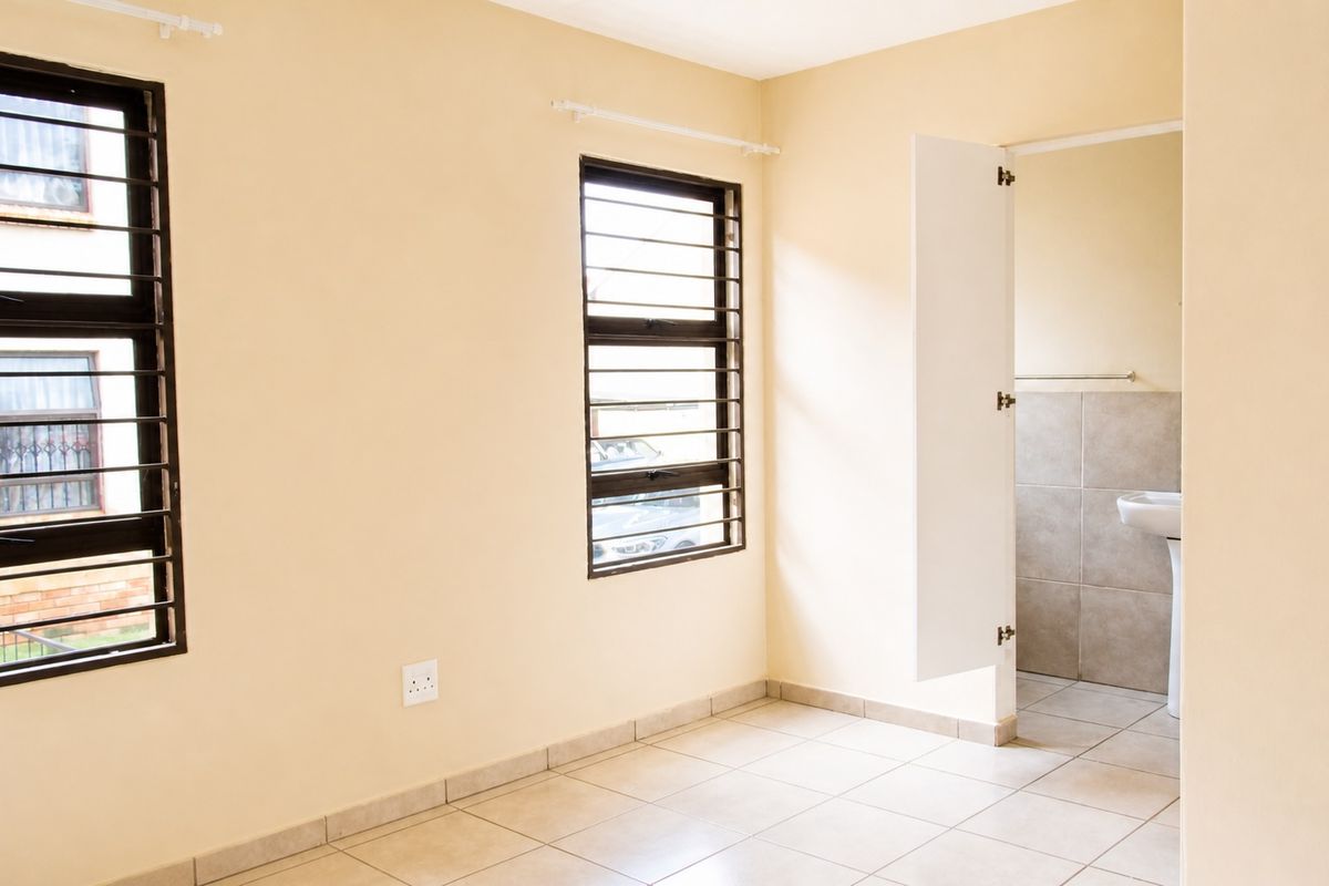 A bright, empty bedroom featuring tiled flooring, security bars on the windows, and an adjoining en-suite bathroom with tiled walls.