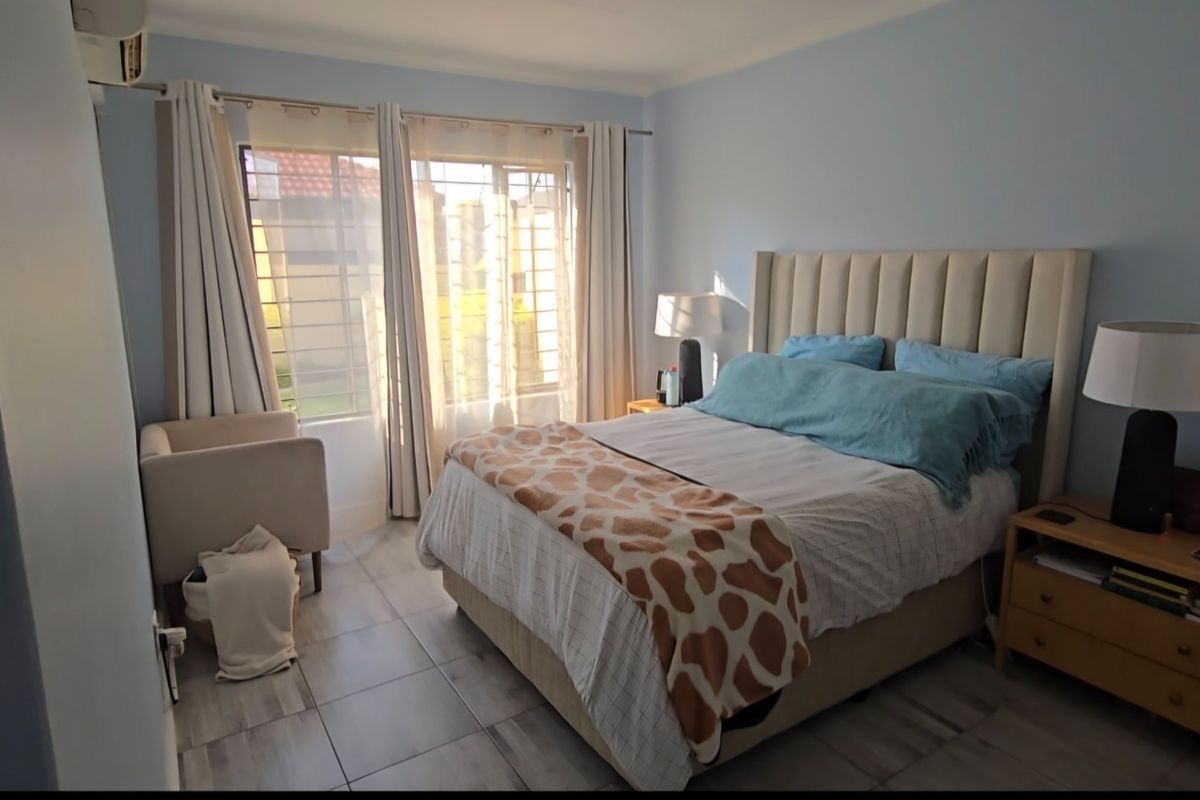 A well-lit bedroom featuring tiled flooring, large windows with security bars, and a wall-mounted air conditioning unit.