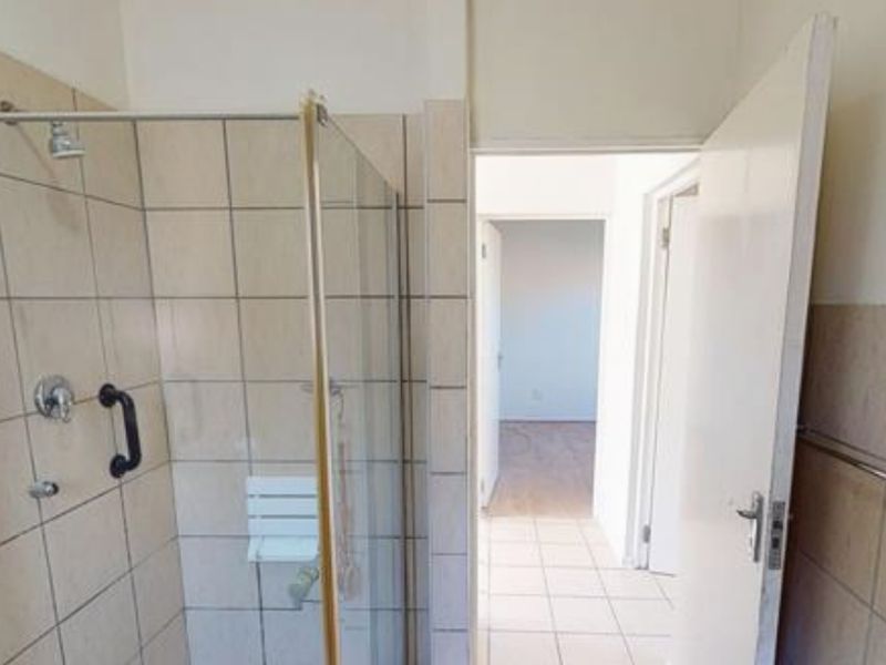 A functional bathroom featuring a shower enclosure with grab bars, tiled walls, and tiled flooring, leading into a hallway.