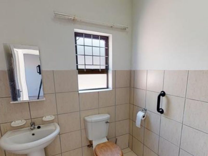 A functional bathroom featuring a pedestal basin, toilet, and wall-mounted grab rail, finished with neutral wall tiling and a window with security bars.