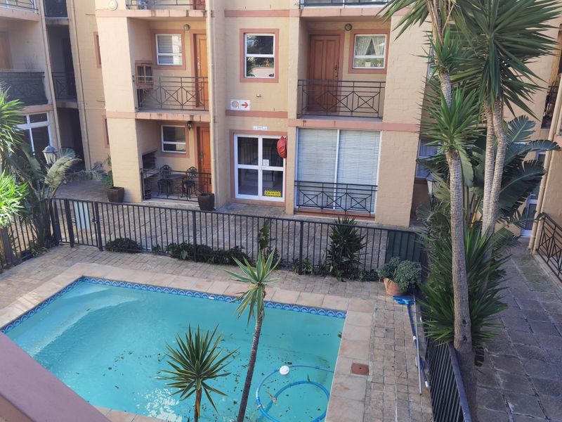 An elevated view of a communal apartment complex pool area featuring paved decking, metal safety fencing, and access to ground-floor balcony units.