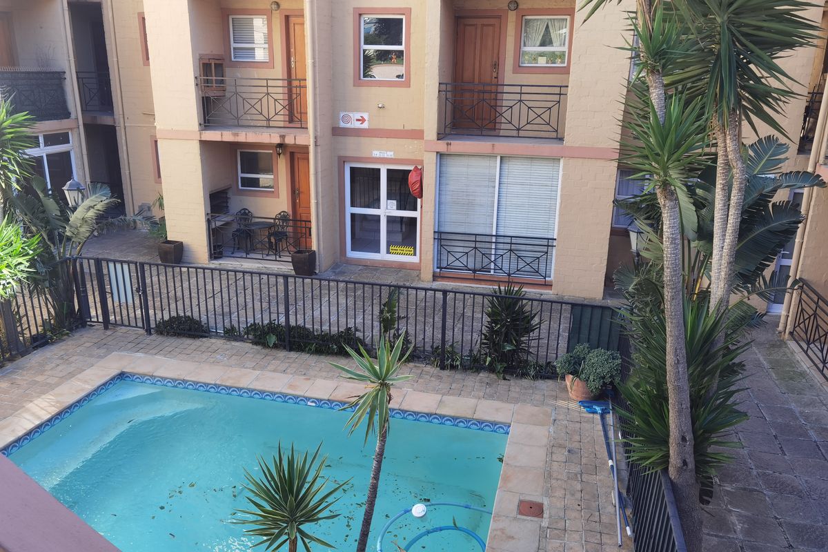 An elevated view of a communal apartment complex pool area featuring paved decking, metal safety fencing, and access to ground-floor balcony units.