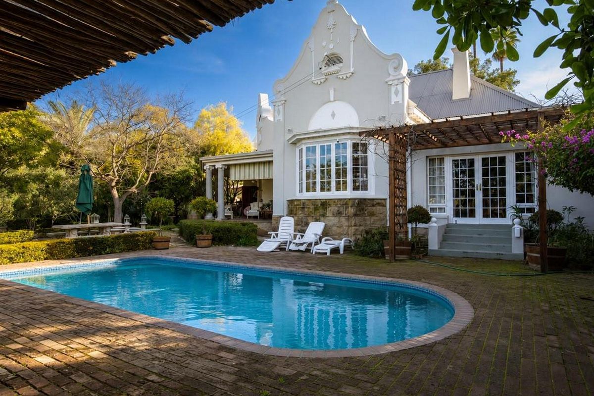 A well-maintained rear garden featuring a swimming pool, brick patio, pergola, and classic Cape Dutch style architecture with french doors.