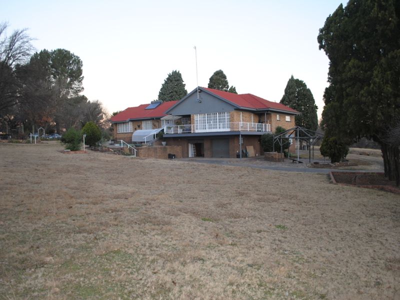 Rear view of a multi-level brick small holding property featuring a red metal roof, raised balcony, and integrated garage set on a large plot.