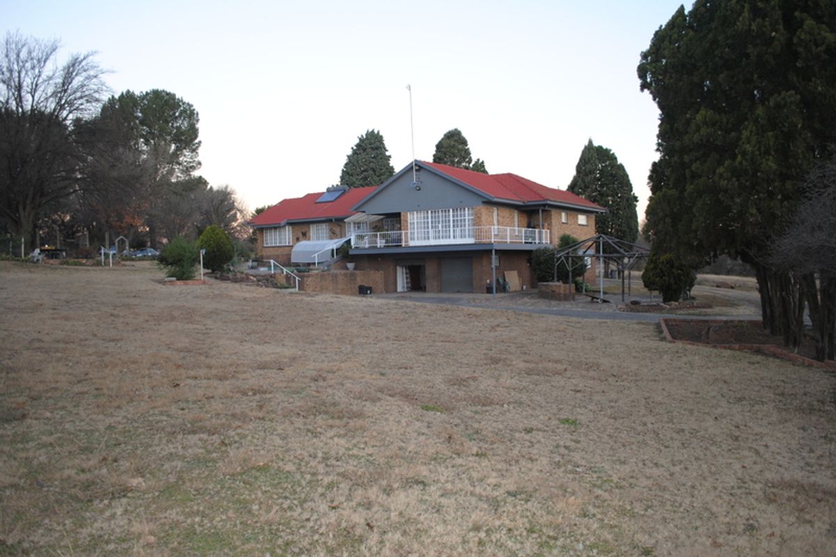 Rear view of a multi-level brick small holding property featuring a red metal roof, raised balcony, and integrated garage set on a large plot.