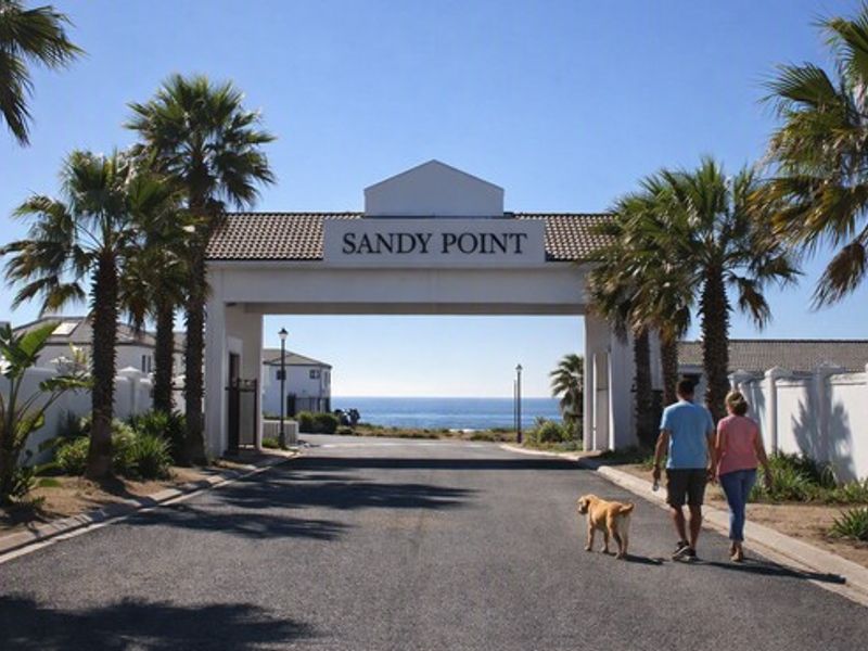 Entrance gate to Sandy Point residential estate featuring paved road access, established palm trees, and ocean views in the distance.