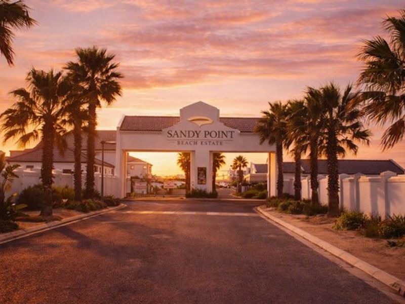 The main entrance gate to Sandy Point Beach Estate, featuring a paved road, perimeter walls, and established palm trees at sunset.