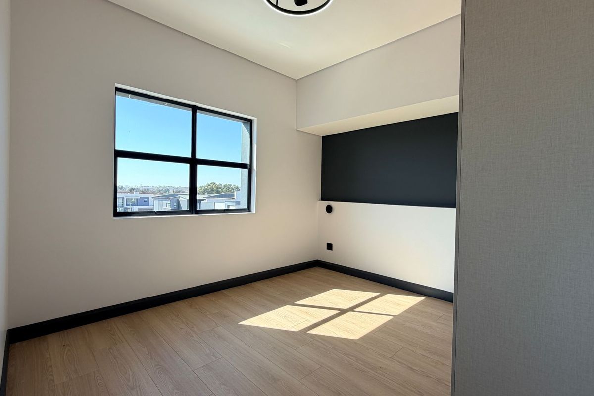 A bright, modern, and empty bedroom featuring light wood-look laminate flooring, a contrasting black accent wall, and a large window with neighborhood views.