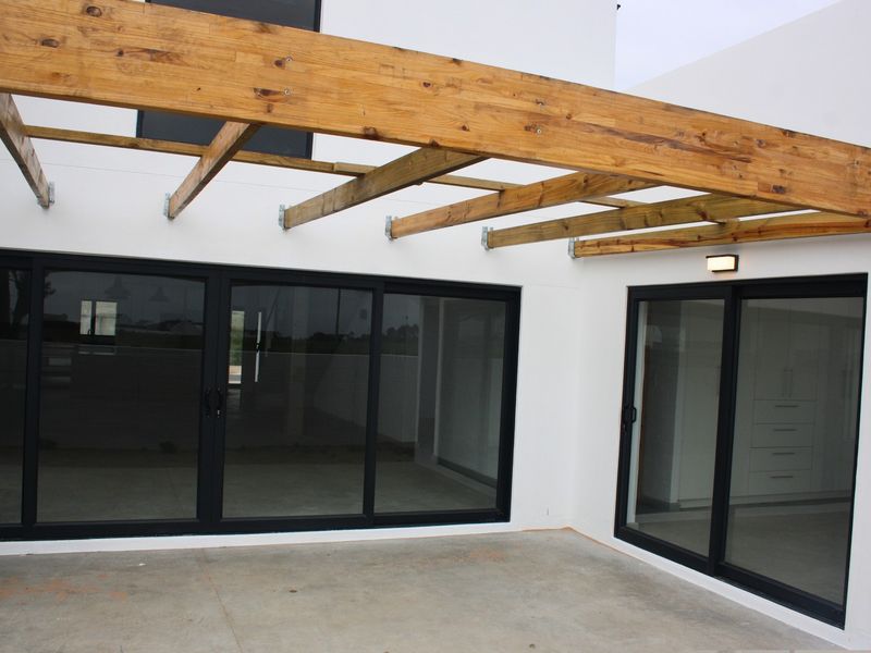 Modern outdoor patio area featuring a timber pergola structure, concrete flooring, and large black-framed sliding glass doors leading into the home.