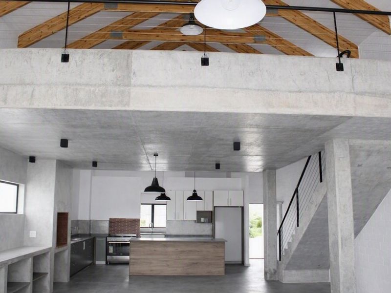 Spacious open-plan living area featuring industrial-style exposed concrete ceilings, timber trusses, a mezzanine level, and a modern kitchen with built-in appliances.