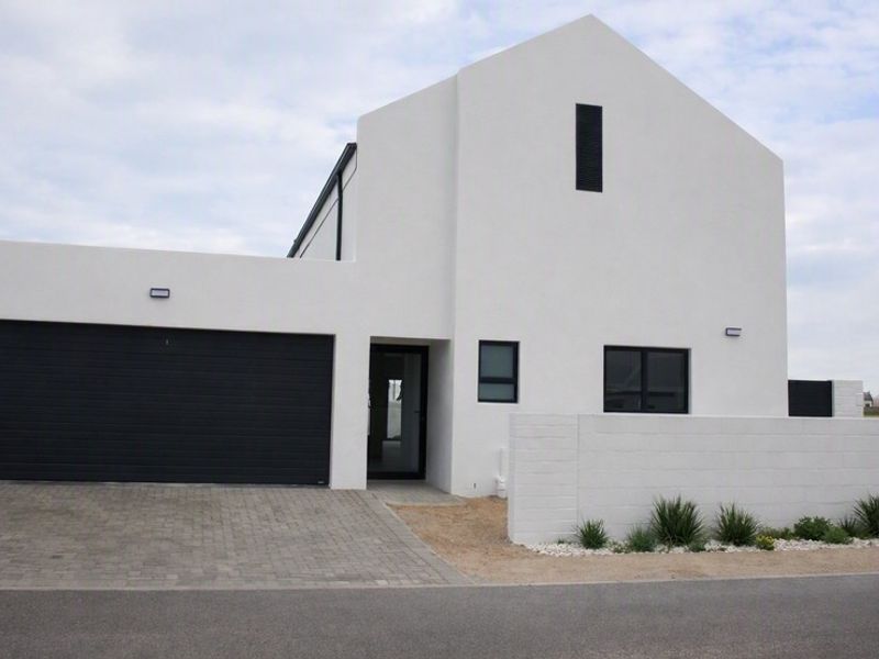 Modern contemporary house exterior featuring a clean white facade, dark garage door, paved driveway, and minimalist boundary wall.