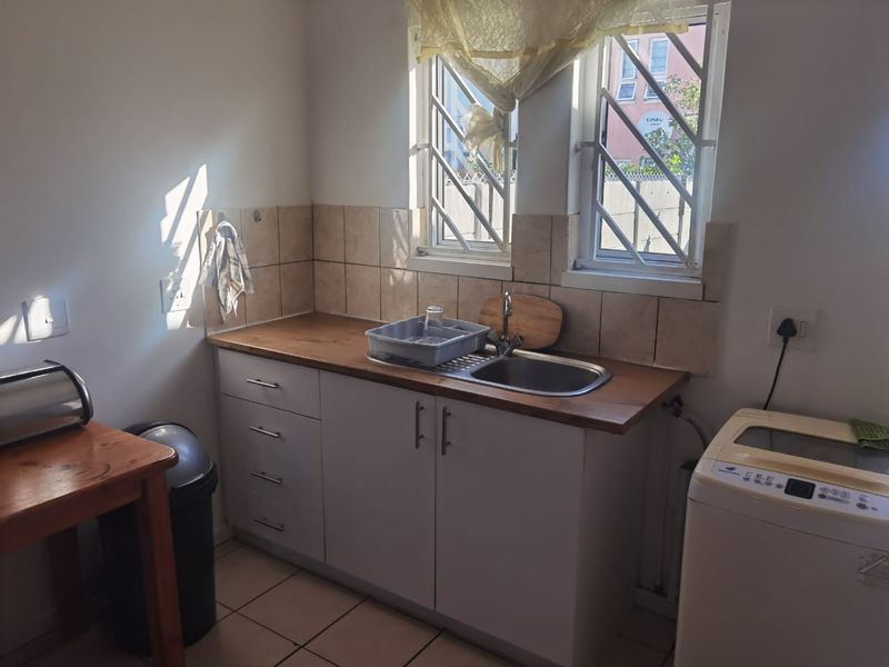 A functional kitchen area featuring a stainless steel sink, tiled backsplash, laminate countertops, and built-in cabinetry with security bars on the windows.