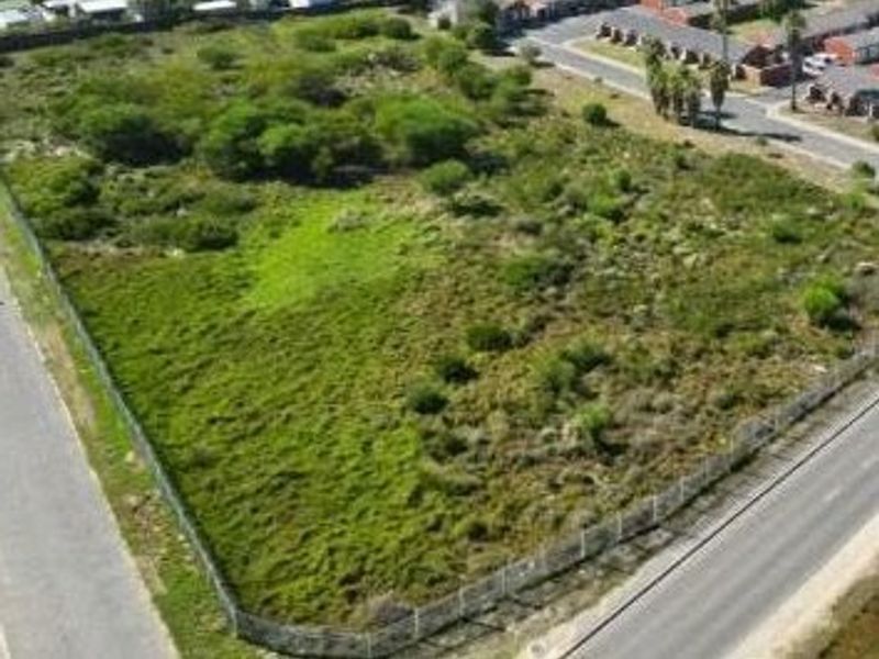 An aerial view of a vacant residential corner plot, featuring perimeter fencing and natural vegetation, situated in a developed neighborhood.