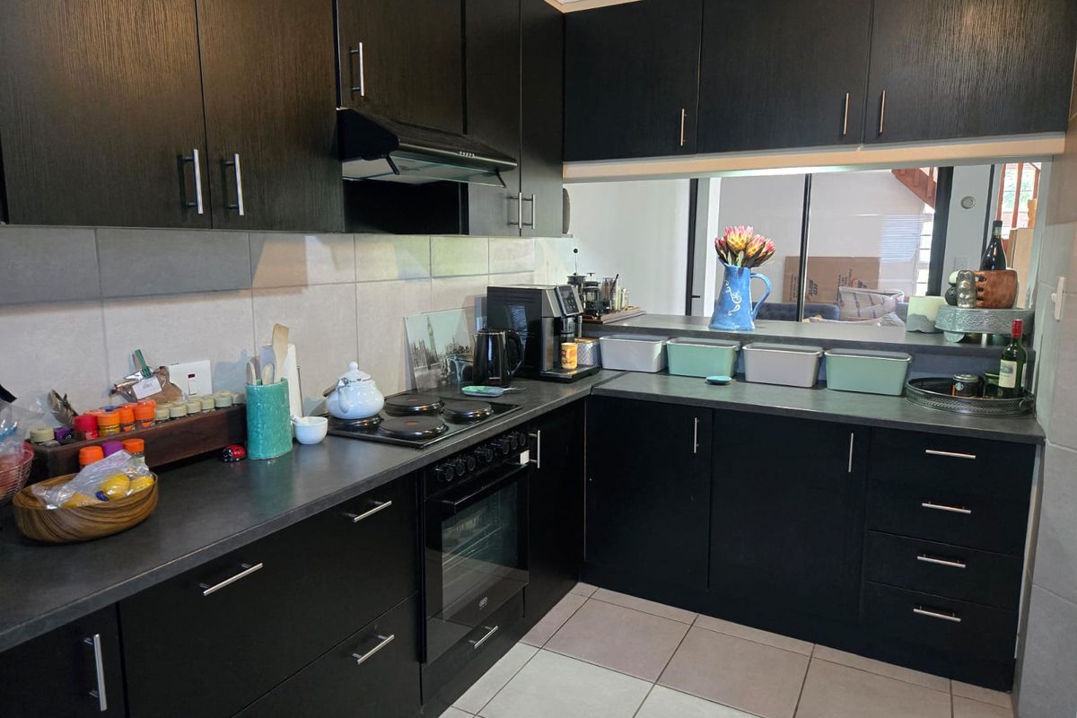 A functional kitchen featuring dark cabinetry, built-in oven and hob, extractor fan, and tiled flooring with a convenient serving hatch to the living area.
