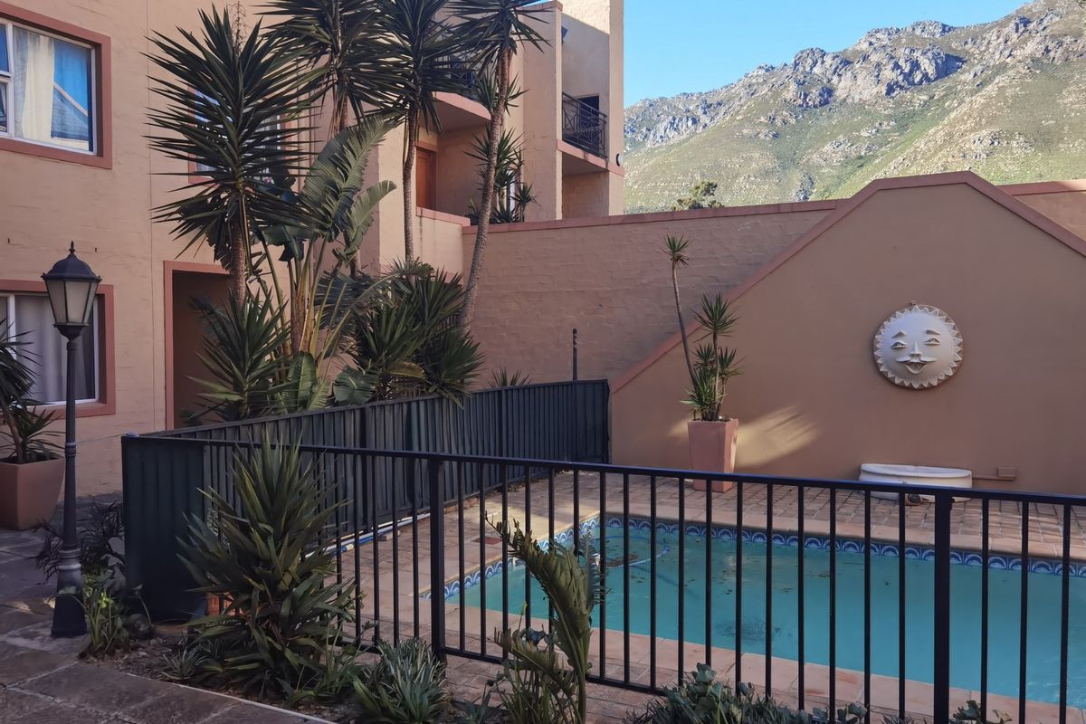 A communal pool area featuring a fenced swimming pool, paved surround, and mountain views, set within an apartment complex courtyard.