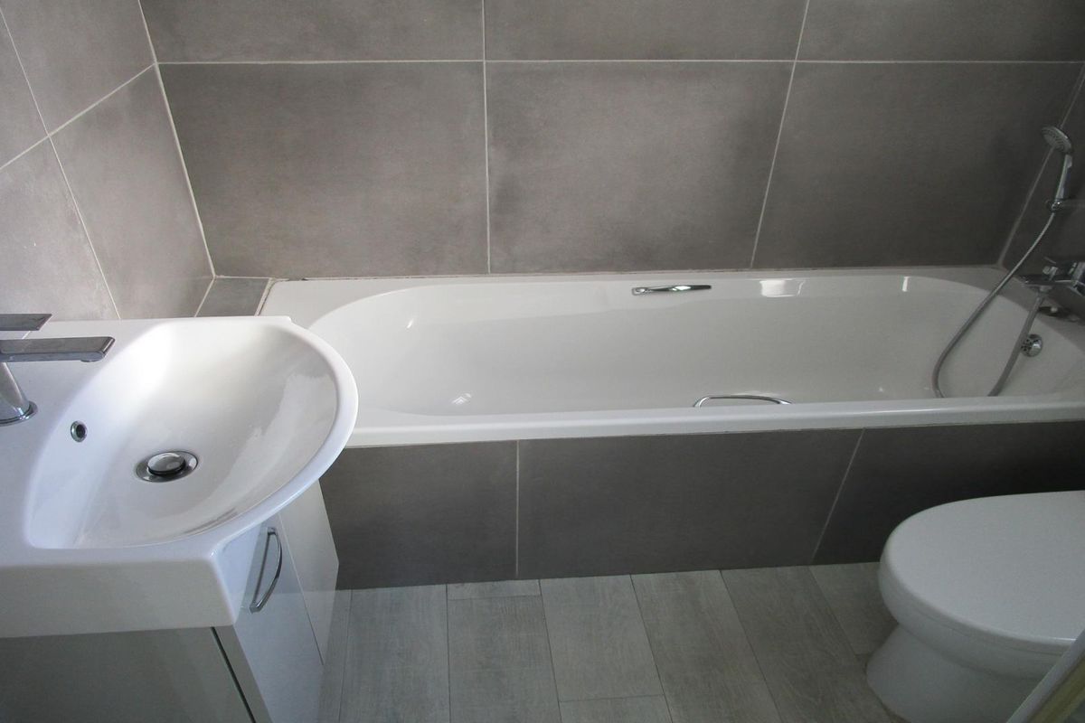 A modern bathroom featuring a built-in bathtub with shower attachment, a vanity basin, and a toilet, finished with neutral grey wall and floor tiles.