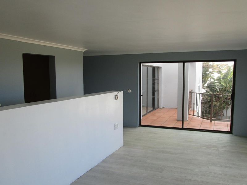 An empty, well-lit living area featuring modern laminate flooring, neutral grey walls, and a sliding door leading to an outdoor balcony.