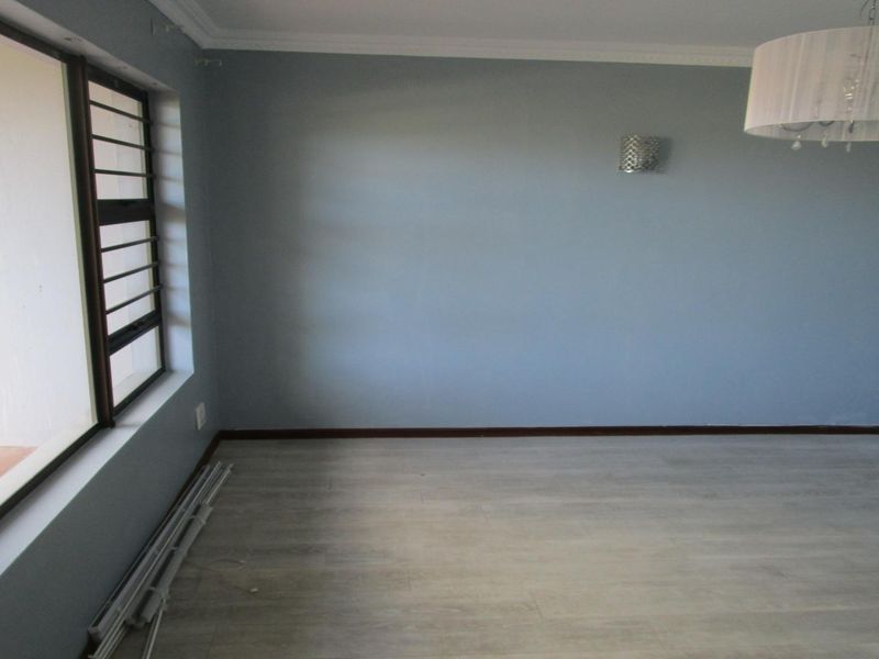 A bright, empty living room featuring light-toned laminate flooring, coved ceilings, and a large window with security bars.