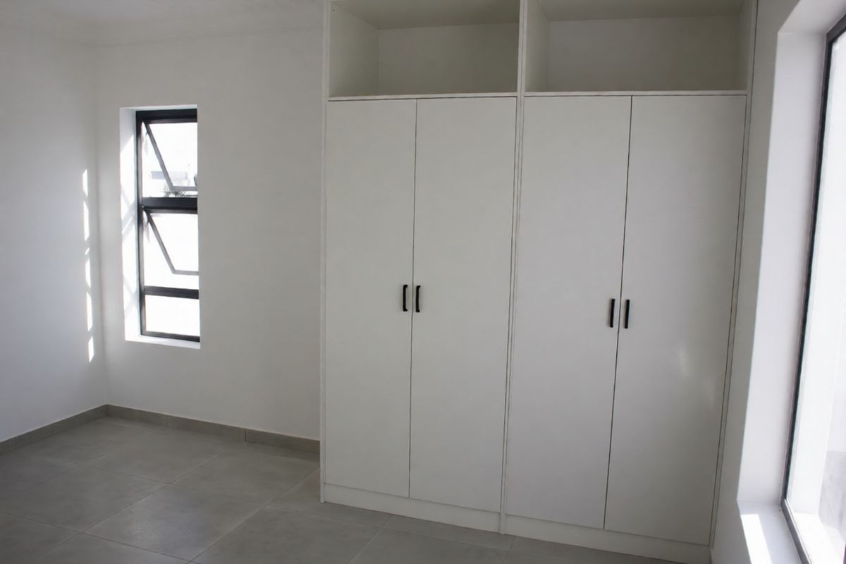A bright, empty bedroom featuring neutral tiled flooring and modern white built-in cupboards with black hardware.
