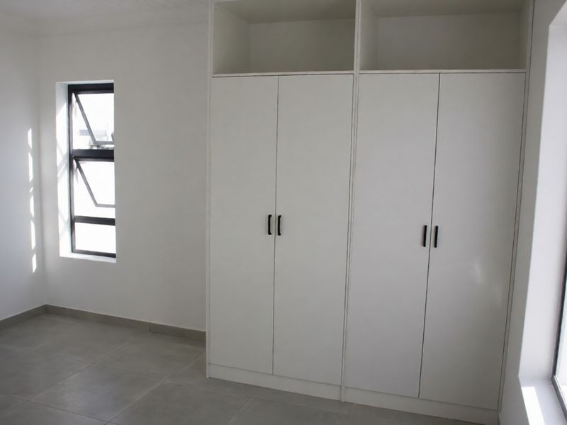 A bright, empty bedroom featuring neutral tiled flooring and modern white built-in cupboards with black hardware.