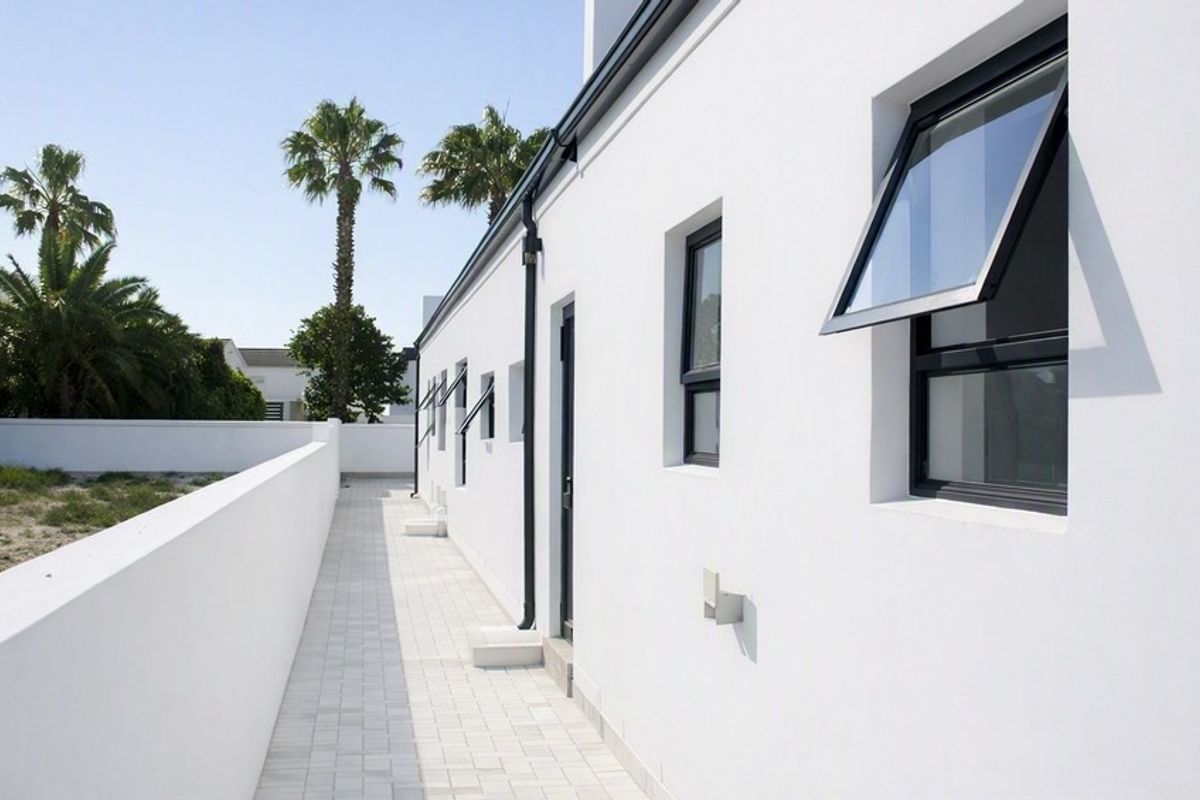 A clean, modern exterior rear walkway featuring white rendered walls, dark-framed casement windows, and paved flooring along the side of the property.
