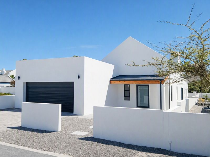 Modern single-story home featuring a clean white facade, dark garage door, and minimalist architectural design with a gravel driveway and boundary wall.