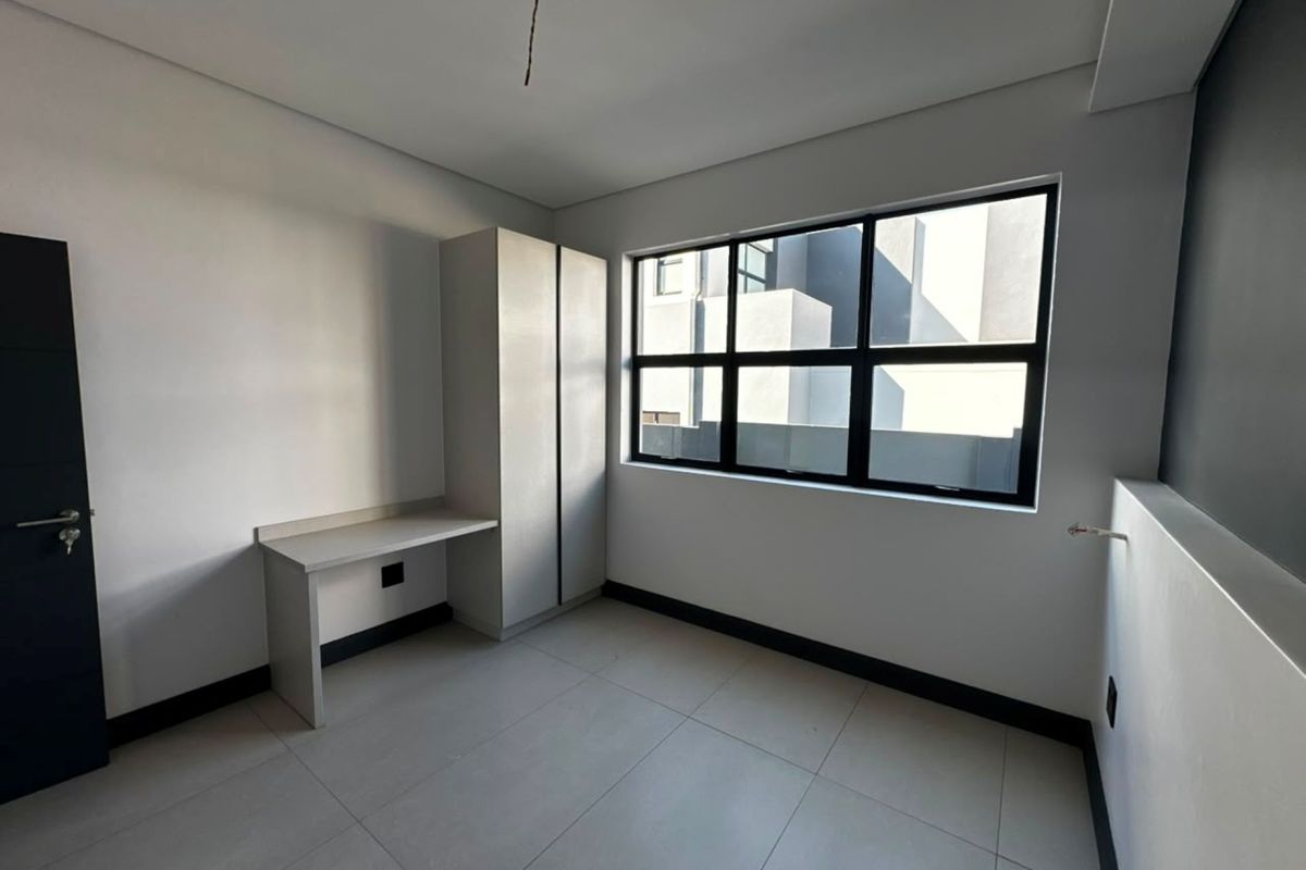 A modern, empty bedroom featuring tiled flooring, a built-in desk, and a built-in wardrobe, with large windows providing natural light.