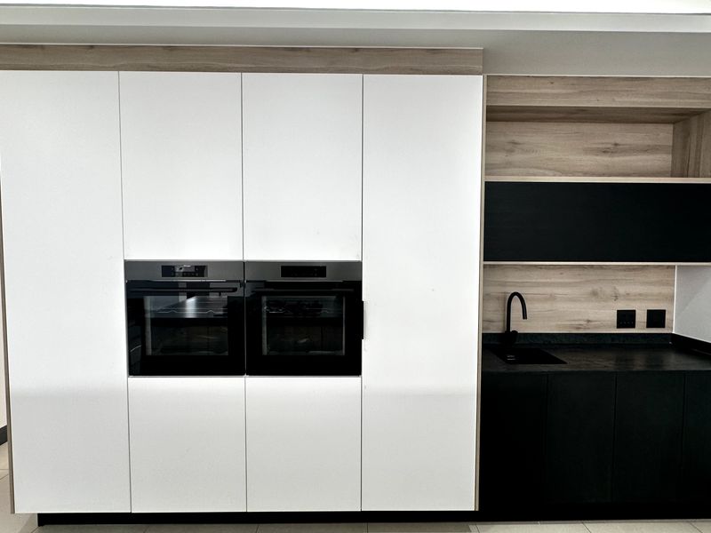Modern kitchen wall featuring integrated double ovens, sleek white and black cabinetry, stone countertops, and a minimalist black sink and tap.