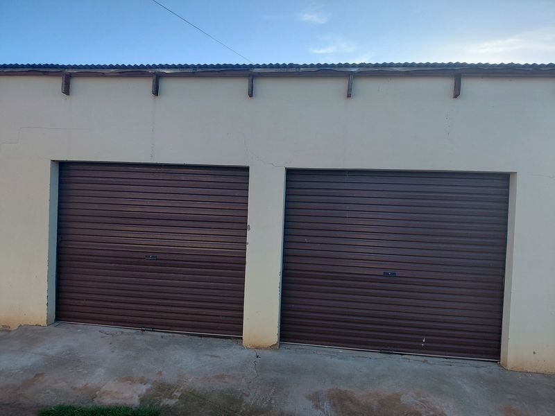 Exterior view of a double garage featuring brown roller doors and a concrete driveway, showing signs of wear on the wall plaster.