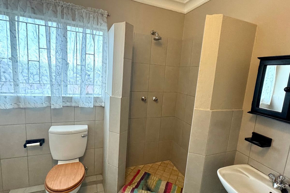 A functional bathroom featuring a tiled shower enclosure, pedestal basin, and toilet, with neutral wall finishes and security bars on the window.