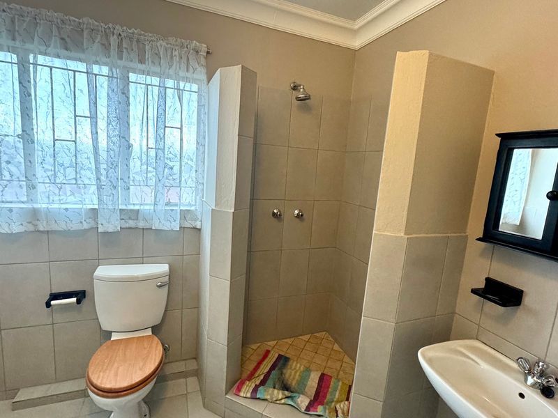 A functional bathroom featuring a tiled shower enclosure, pedestal basin, and toilet, with neutral wall finishes and security bars on the window.
