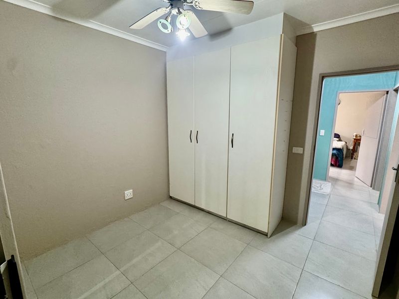 A clean, empty bedroom featuring neutral wall tones, tiled flooring, and built-in white cupboards with a ceiling fan.