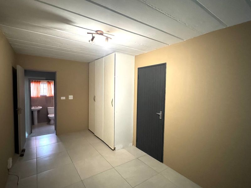 A simple bedroom featuring tiled flooring, built-in cupboards, and an exposed beam ceiling, with an adjoining bathroom visible through the doorway.