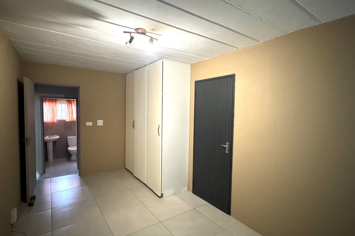 A simple bedroom featuring tiled flooring, built-in cupboards, and an exposed beam ceiling, with an adjoining bathroom visible through the doorway.