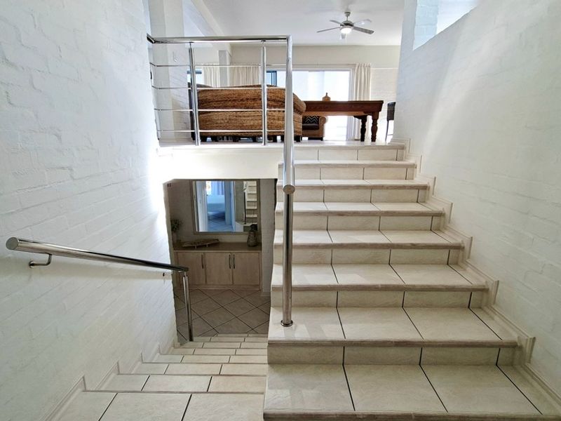 A bright split-level hallway featuring tiled stairs, stainless steel railings, and painted brick walls, connecting different living areas of the home.
