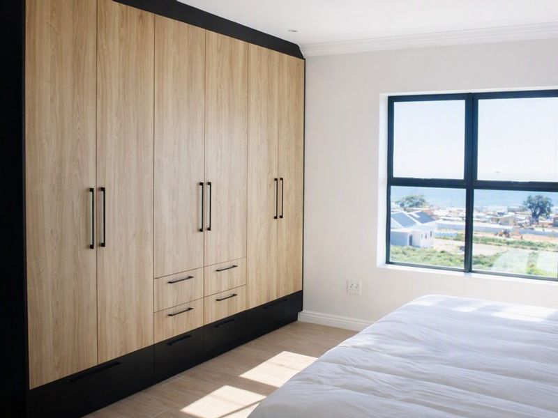 A modern bedroom featuring extensive built-in wooden cupboards with black accents and a window offering a coastal view.