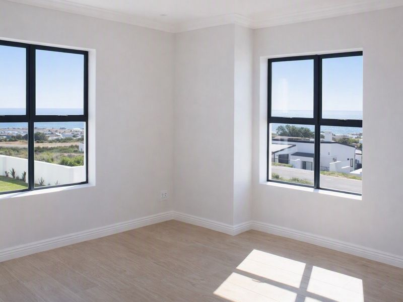 A bright, empty bedroom featuring modern laminate flooring, crisp white walls, and large windows offering scenic coastal views.