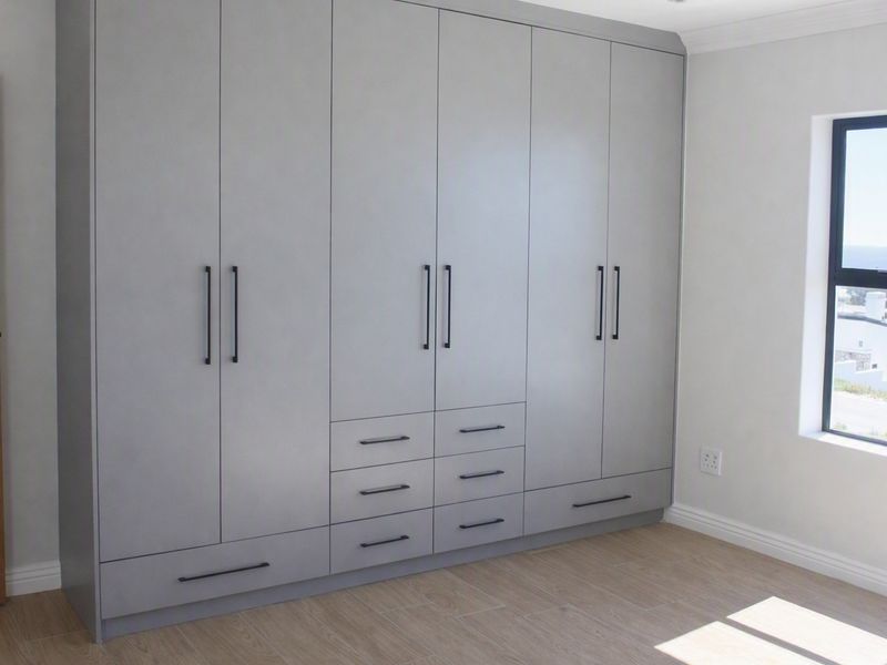A bright bedroom featuring modern grey built-in cupboards, light wood-look laminate flooring, and a window with coastal views.