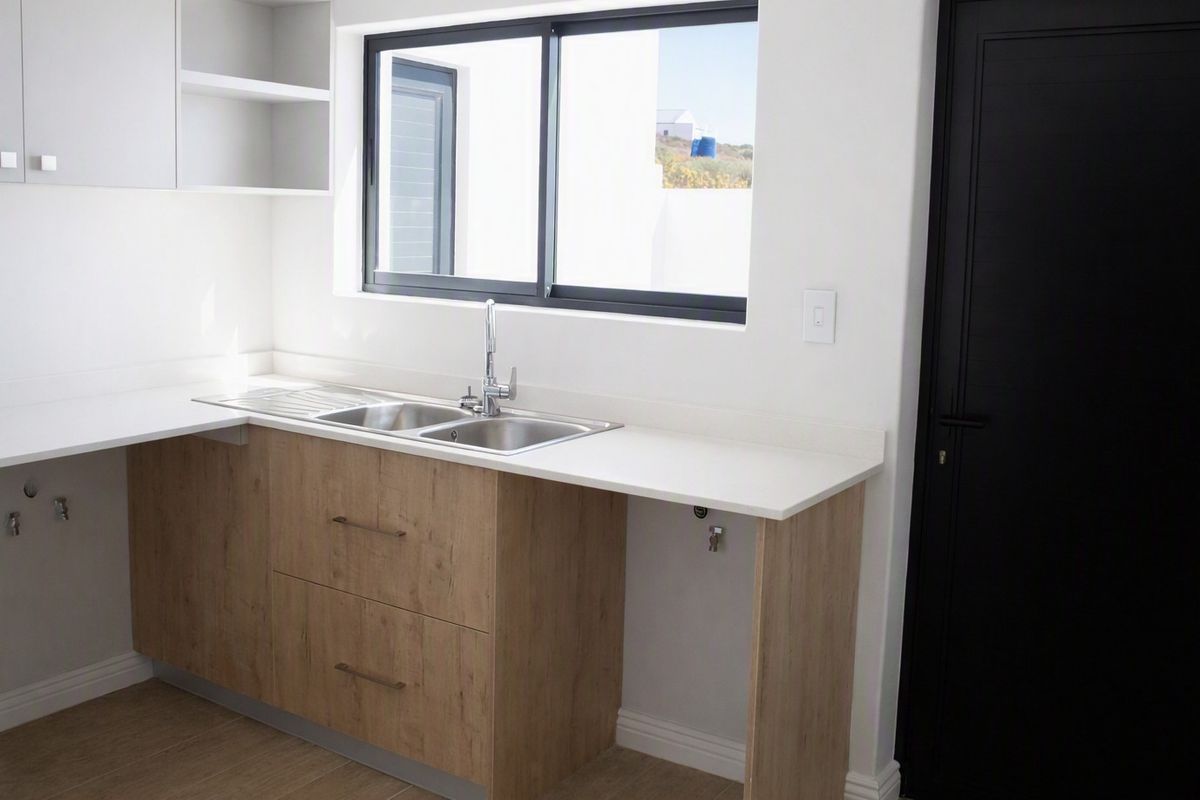A modern, clean kitchen area featuring light wood-finish cabinetry, a stainless steel sink, and a bright window, presented in excellent condition.