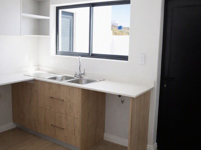 A modern, clean kitchen area featuring light wood-finish cabinetry, a stainless steel sink, and a bright window, presented in excellent condition.