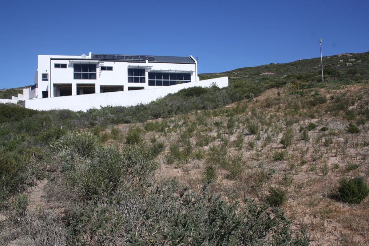 A modern multi-level residential property situated on a sloped plot, featuring large windows, solar panels, and a prominent white retaining wall.
