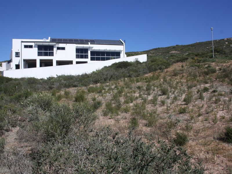 A modern multi-level residential property situated on a sloped plot, featuring large windows, solar panels, and a prominent white retaining wall.