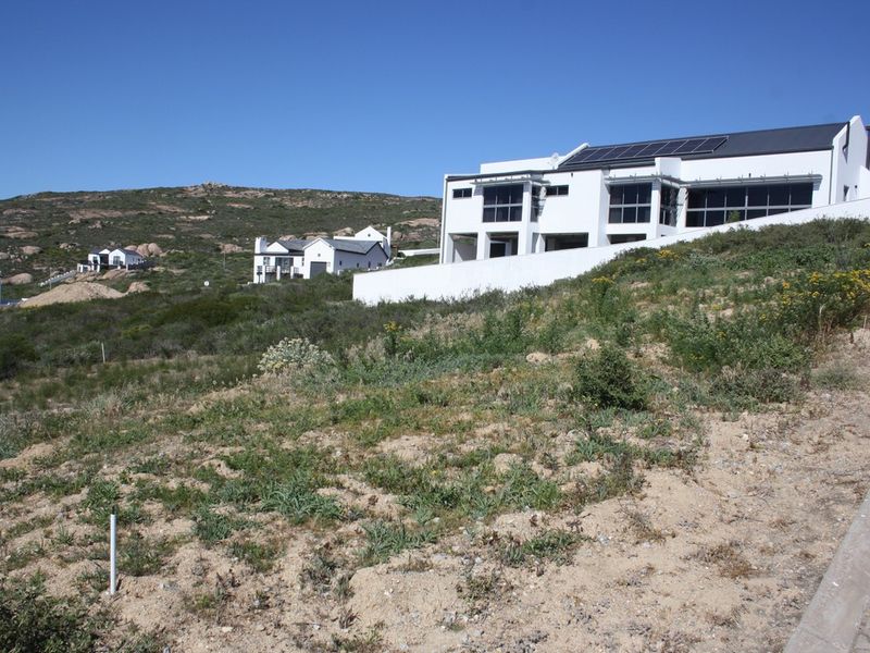 A view of a vacant residential plot with a sloping landscape, featuring a modern multi-story home in the background with solar panels.