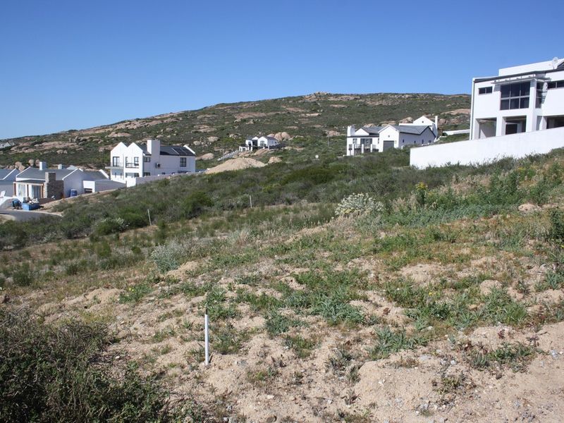 A sloped, undeveloped residential plot in a coastal estate, featuring natural vegetation and surrounding modern residential homes.