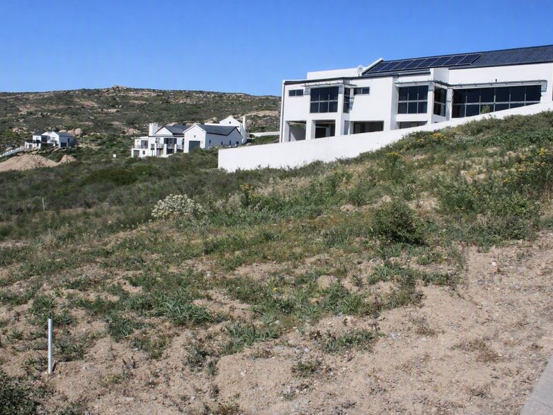 A sloped residential vacant land plot with natural vegetation, situated in a developed estate with neighbouring modern homes.