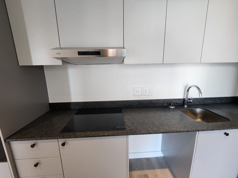 A modern, compact kitchen featuring white cabinetry, dark stone countertops, an integrated induction hob, stainless steel extractor fan, and a sleek sink.