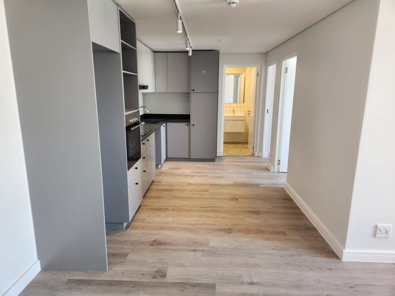 Modern open-plan kitchen featuring sleek grey and white cabinetry, stone countertops, built-in oven, and light wood-look laminate flooring.