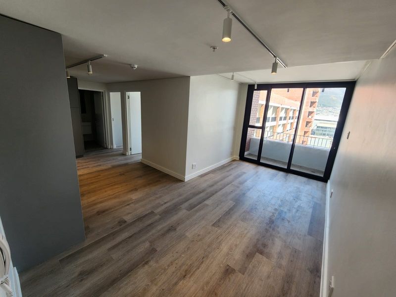 A bright, empty living area featuring modern laminate flooring, track lighting, and large sliding glass doors leading to a private balcony.