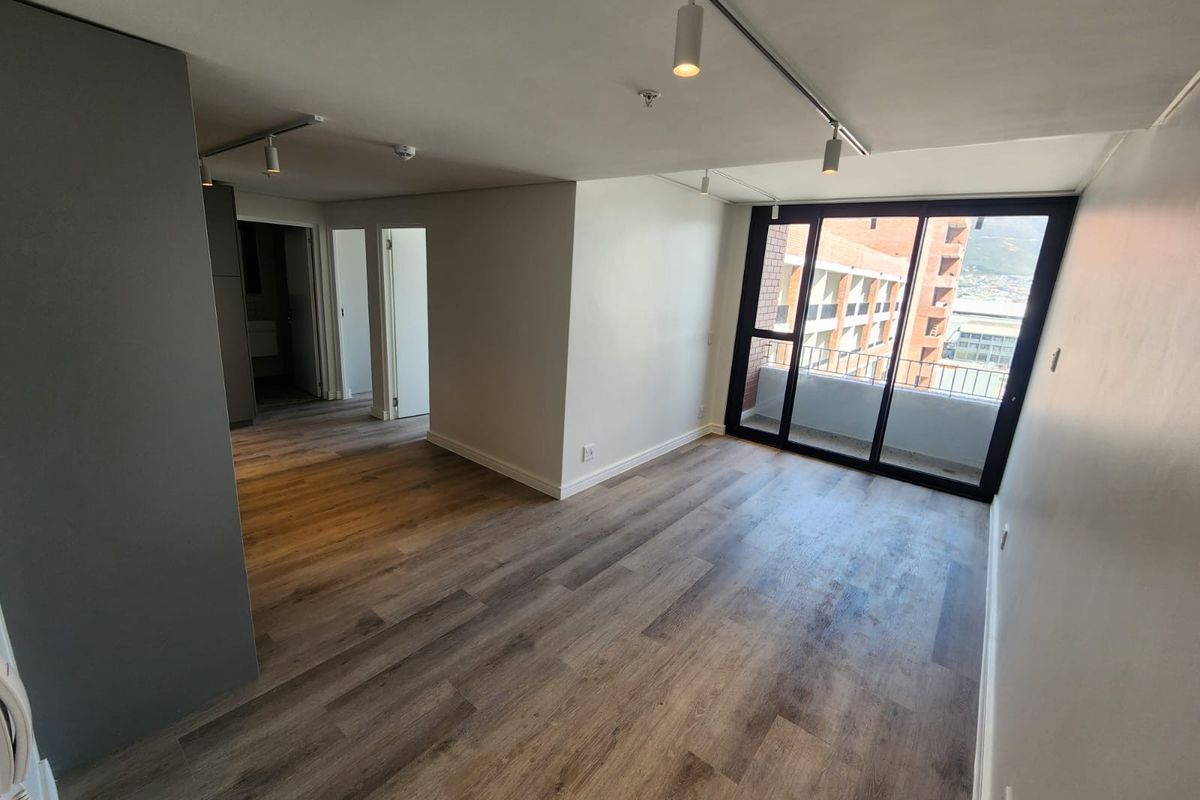 A bright, empty living area featuring modern laminate flooring, track lighting, and large sliding glass doors leading to a private balcony.