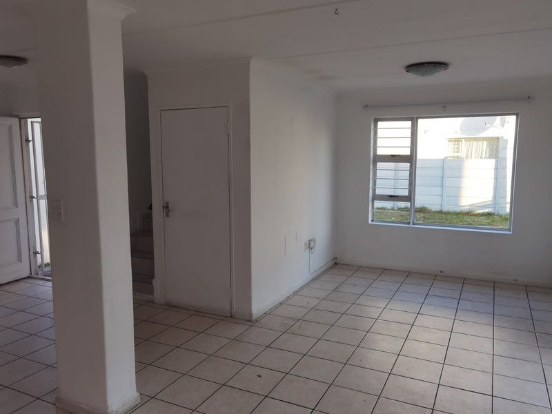 An empty, neutrally painted living area featuring tiled flooring, a large window with security bars, and direct access to an adjacent space.